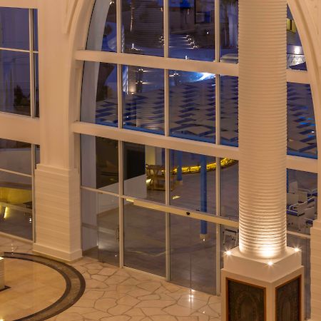 Pickalbatros Palace Sharm - "Aqua Park" Exterior photo The photo shows a spacious interior of a modern building, likely a hotel or a lobby. The scene features large glass windows that allow for a view of areas outside. There are elegant pillars within the space, illuminated from the base, creating a warm