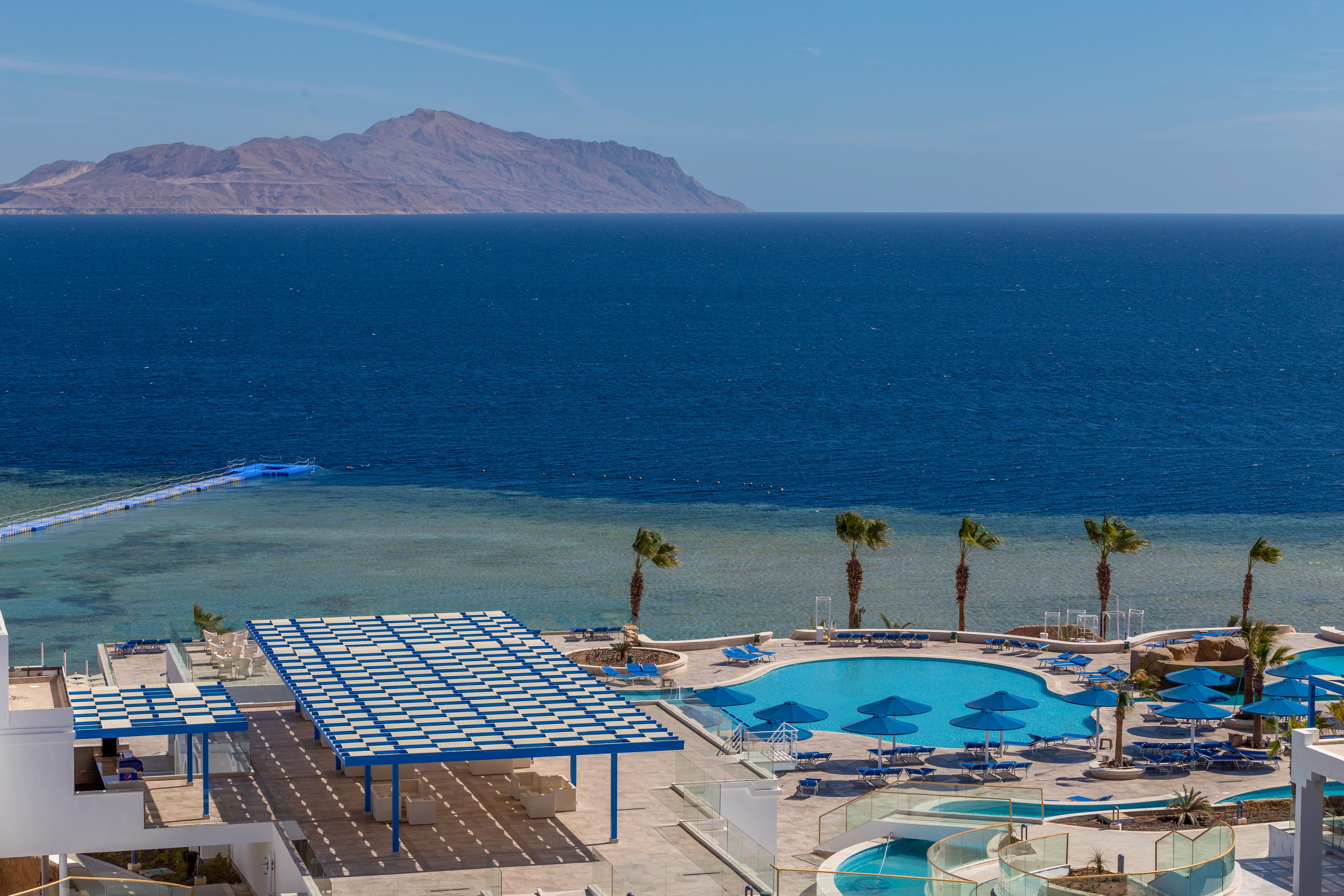 Pickalbatros Palace Sharm - "Aqua Park" Exterior photo The photo shows a beautiful coastal scene featuring a large swimming pool area with lounge chairs and umbrellas. In the background, there is a clear blue ocean stretching out to the horizon, with a distant landmass or mountain visible. Palm trees are