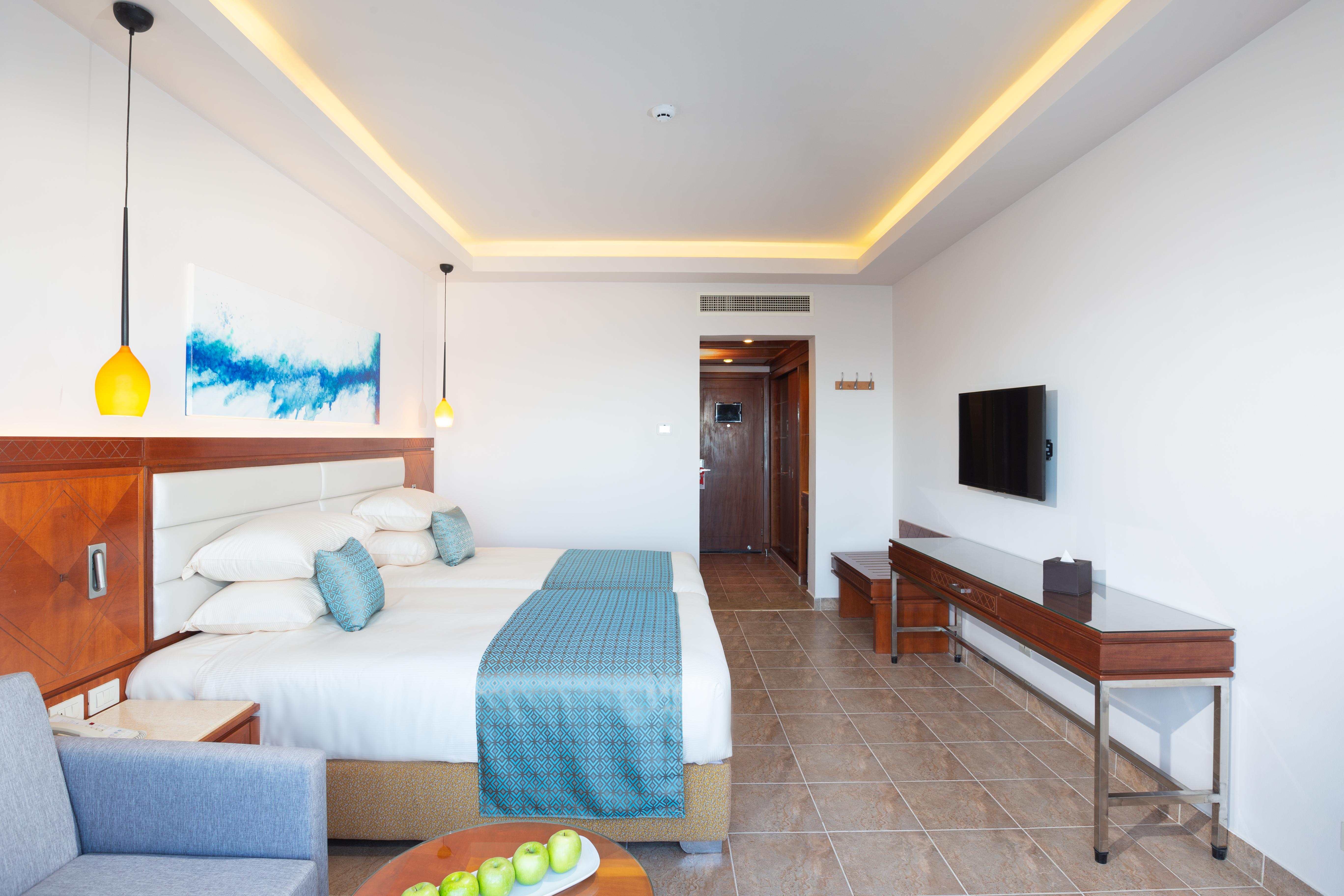 Pickalbatros Palace Sharm - "Aqua Park" Exterior photo The photo shows a modern hotel room featuring two queen-sized beds with white linens and a light blue throw. The room is well-lit, with a warm yellow glow from the ceiling lights. There is a painting on the wall above the beds, characterized by blue 