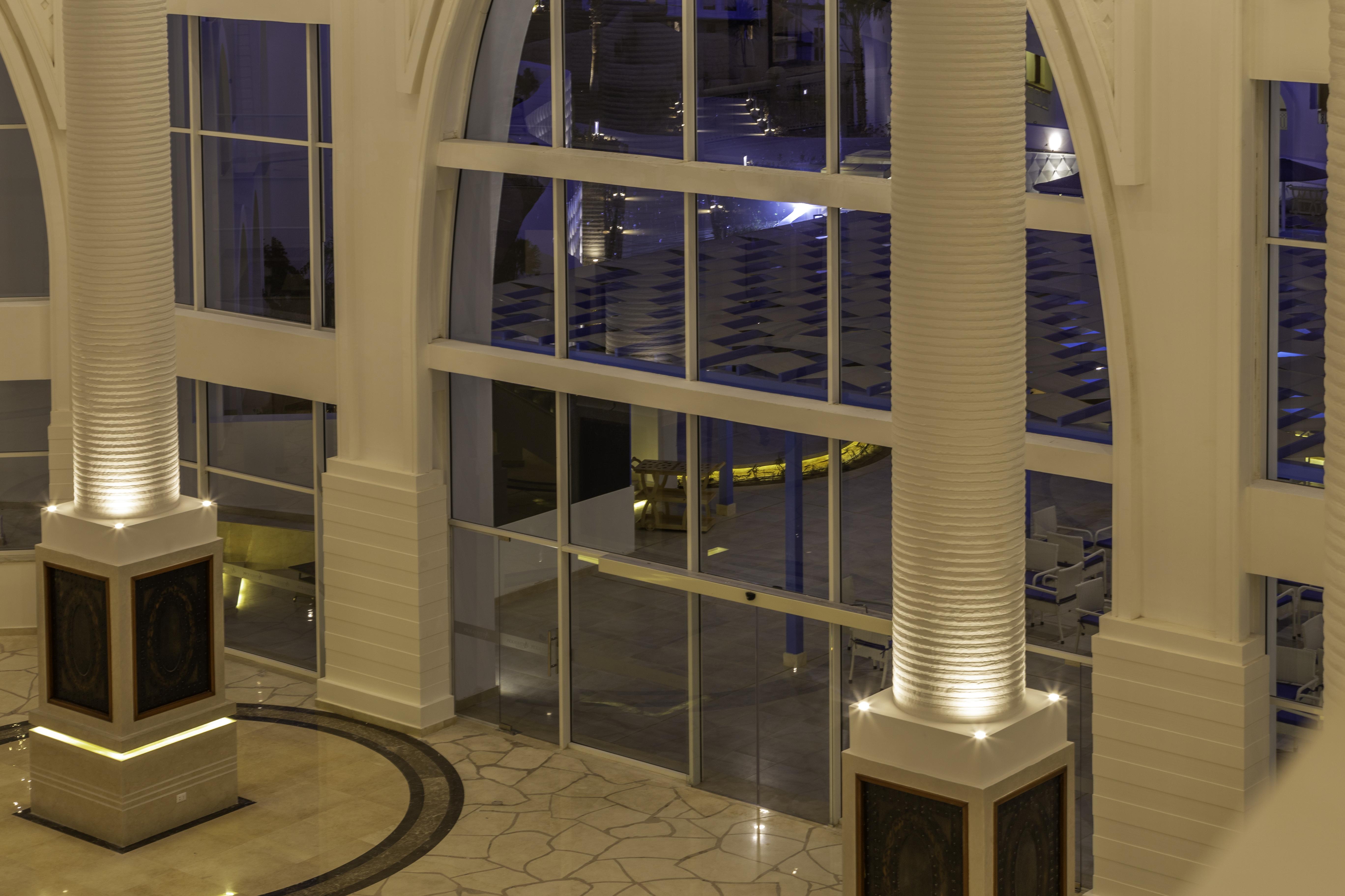 Pickalbatros Palace Sharm - "Aqua Park" Exterior photo The photo shows a spacious interior of a modern building, likely a hotel or a lobby. The scene features large glass windows that allow for a view of areas outside. There are elegant pillars within the space, illuminated from the base, creating a warm