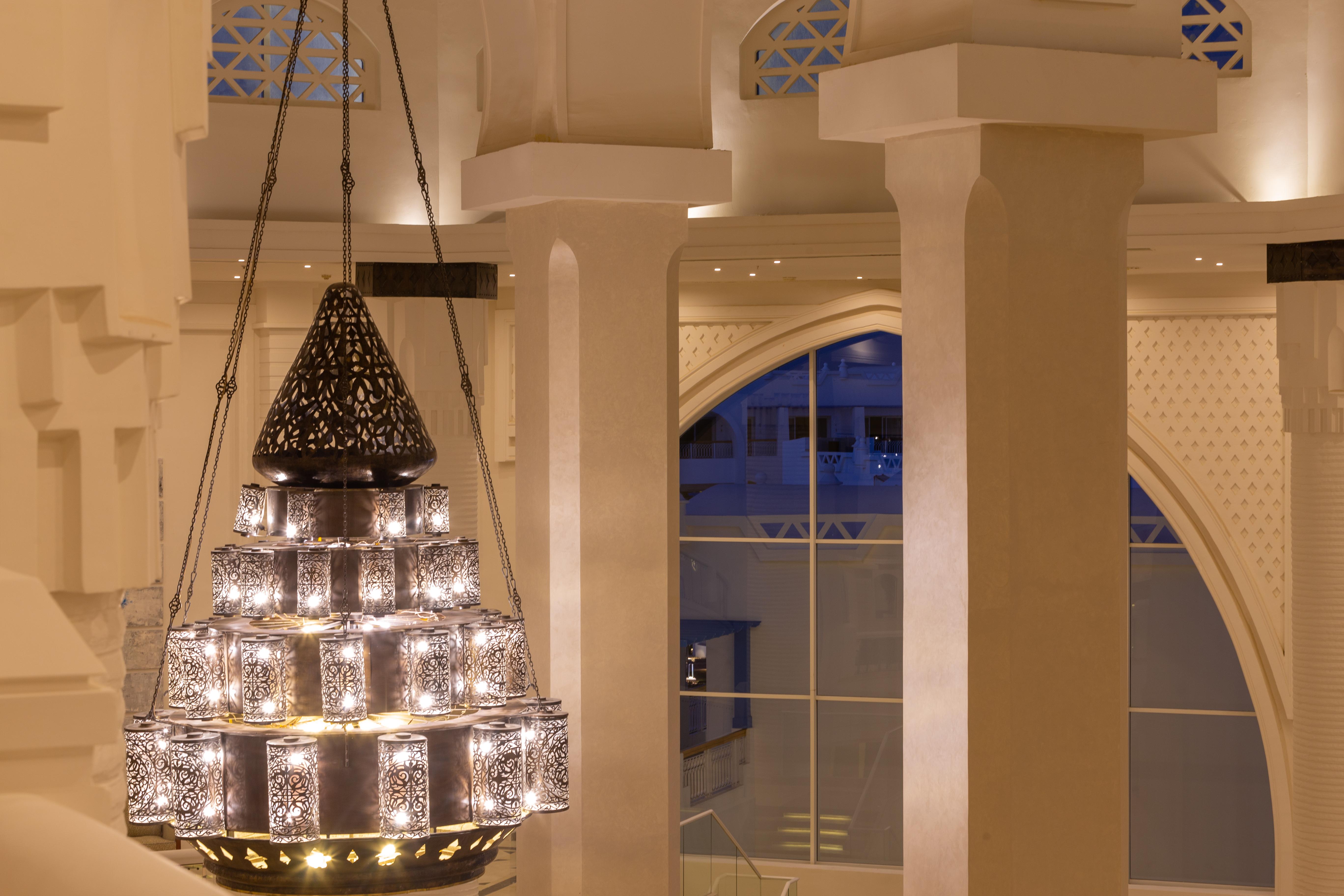 Pickalbatros Palace Sharm - "Aqua Park" Exterior photo The photo shows a beautifully designed chandelier hanging from the ceiling of a spacious interior. The chandelier features a structure with multiple levels and includes ornate patterns, likely made of metal or glass, that diffuse light elegantly. The