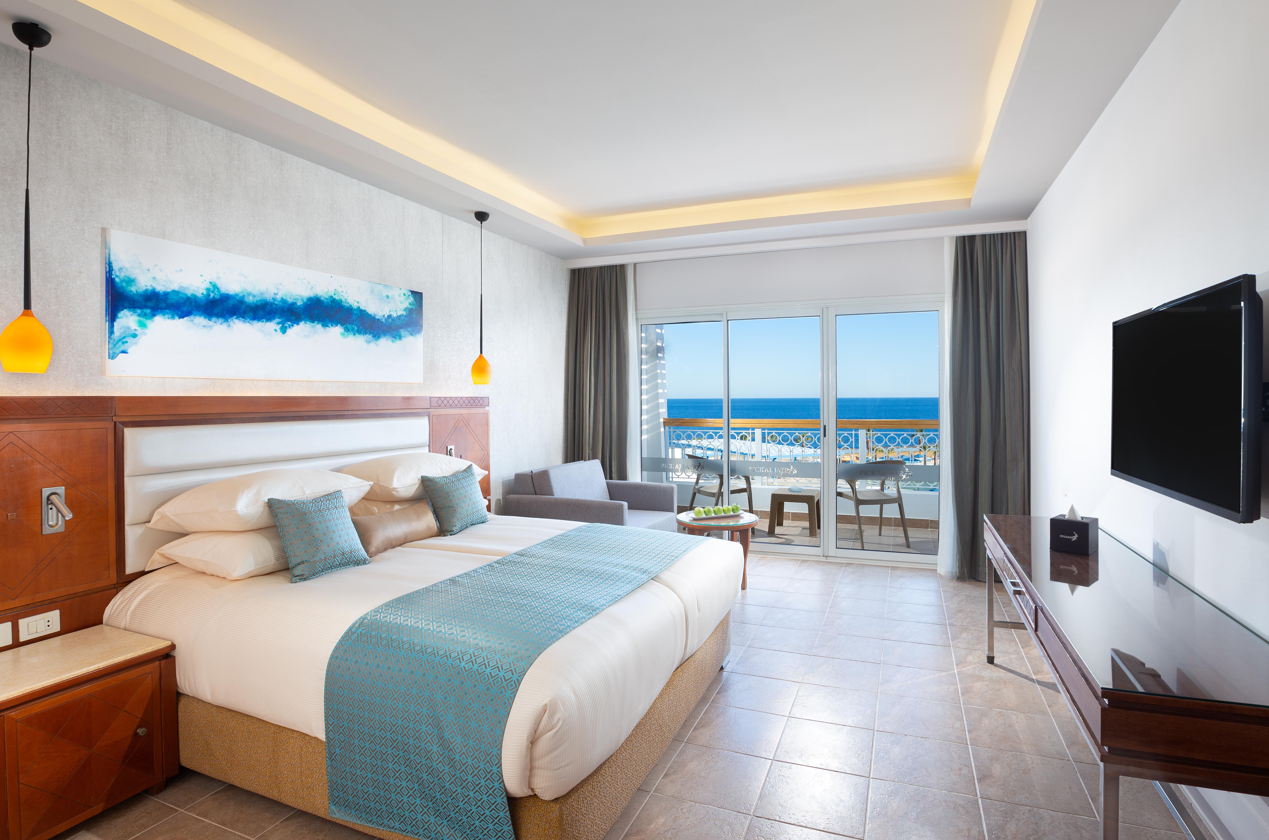 Pickalbatros Palace Sharm - "Aqua Park" Exterior photo The photo shows a modern hotel room with a large bed dressed in white linens and a light blue throw blanket. There is a piece of abstract artwork on the wall above the bed, featuring blue tones. The room has large windows with a view of the ocean, al