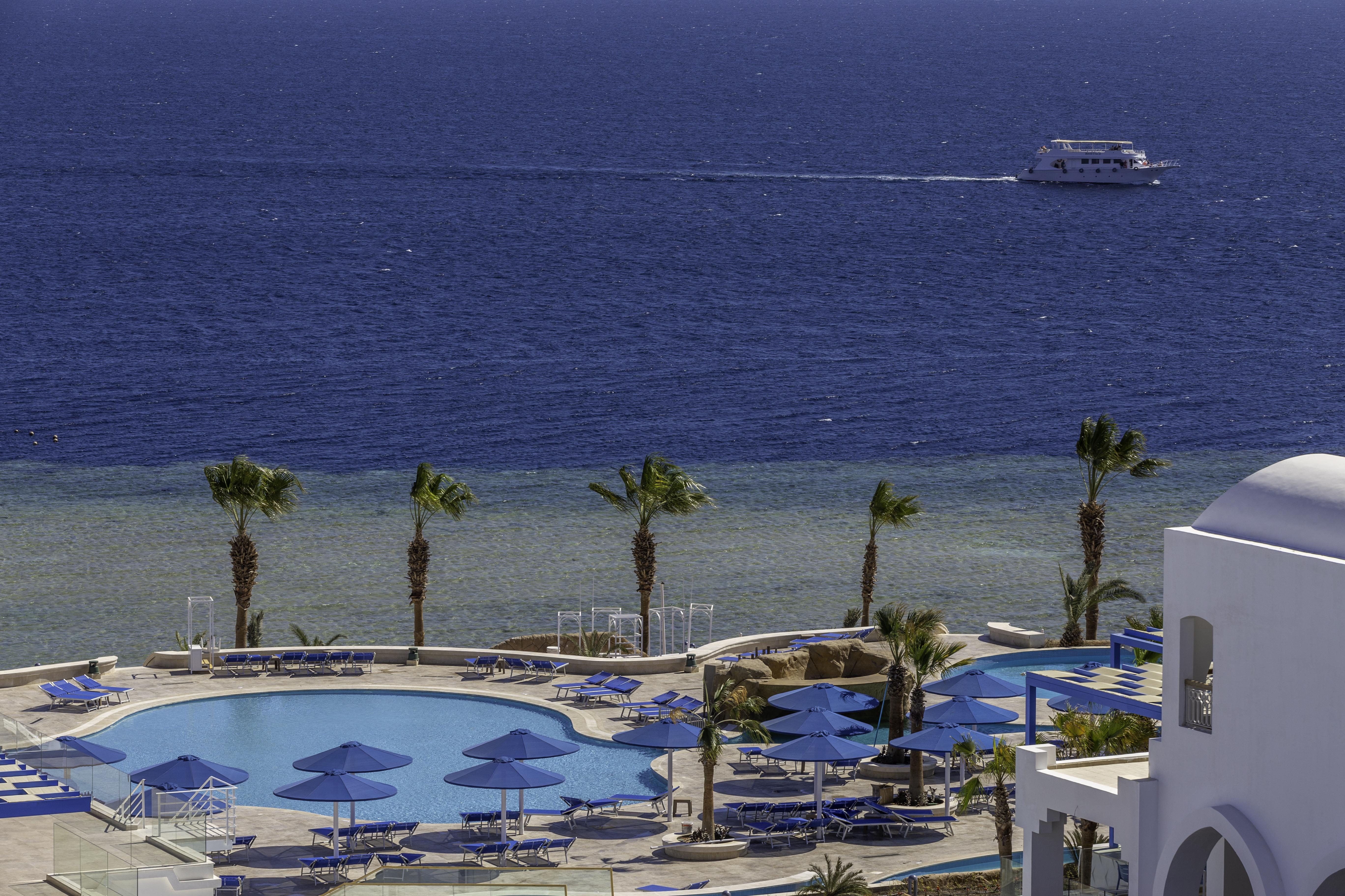 Pickalbatros Palace Sharm - "Aqua Park" Exterior photo The photo depicts a serene seaside view featuring a large swimming pool surrounded by lounge chairs and umbrellas. In the background, there is a calm sea with a boat cruising on the water. Palm trees line the edge of the pool area, adding to the trop