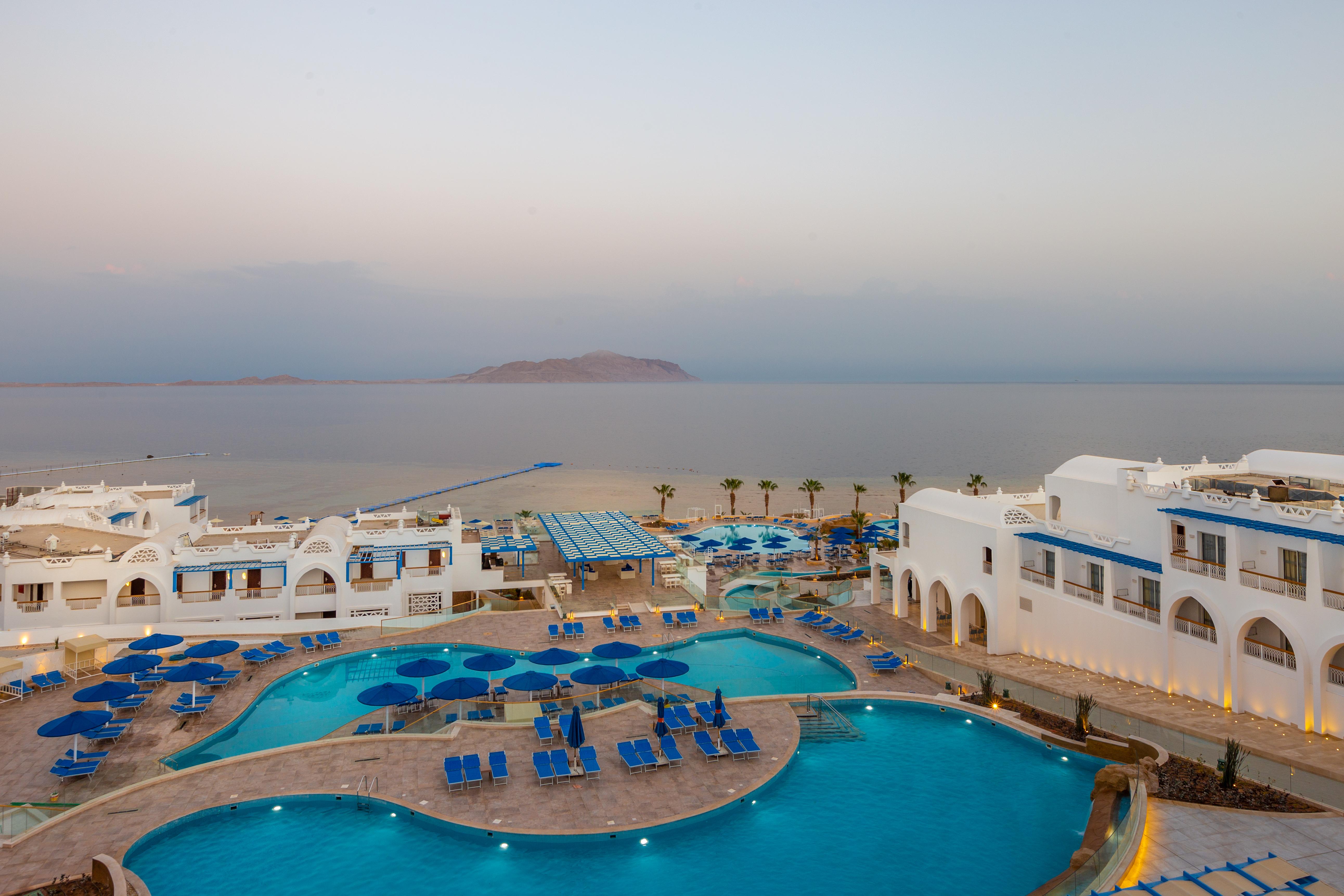 Pickalbatros Palace Sharm - "Aqua Park" Exterior photo The photo shows a resort area with expansive pools surrounded by lounge chairs. The pools are designed in a curvy shape, and there are numerous blue umbrellas scattered around. In the background, there is a calm sea stretching out towards a distant l