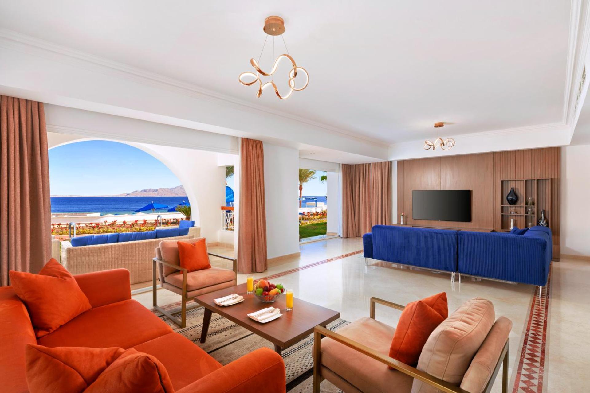 Pickalbatros Palace Sharm - "Aqua Park" Exterior photo The photo showcases a stylish and modern living space that features a bright and open layout. The room includes a cozy seating area with orange couches and a wooden coffee table, adorned with a fruit arrangement. Large windows provide a view of the o