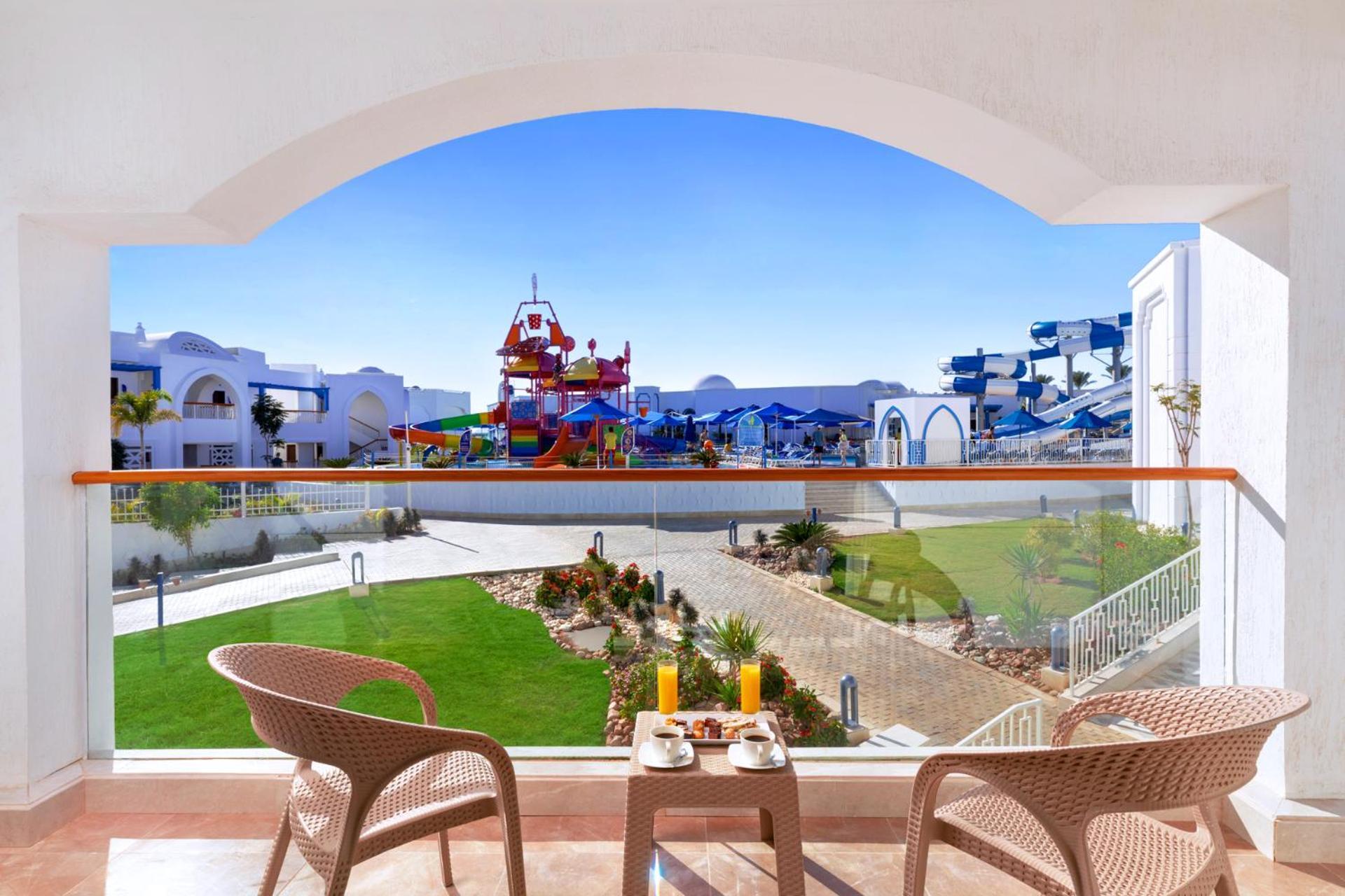 Pickalbatros Palace Sharm - "Aqua Park" Exterior photo The photo shows a view from a balcony with two chairs and a small table set up for relaxation. In the foreground, there is a breakfast setup with orange juice and a pastry. Beyond the balcony, there is a vibrant, colorful water play area with slides 
