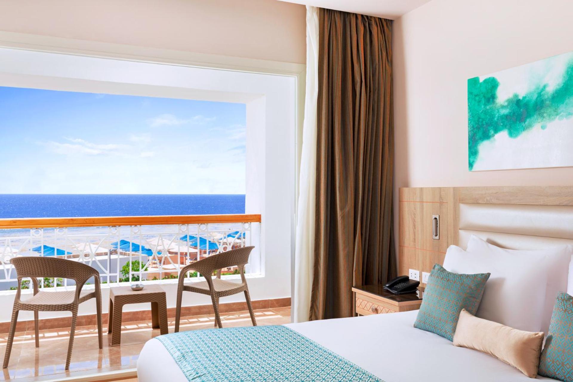 Pickalbatros Palace Sharm - "Aqua Park" Exterior photo The photo shows a modern hotel room featuring a large bed with white linens and a teal accent pillow. There is a window with a view of the ocean, framed by light curtains, which lets in natural light. In front of the window, there are two wooden chai