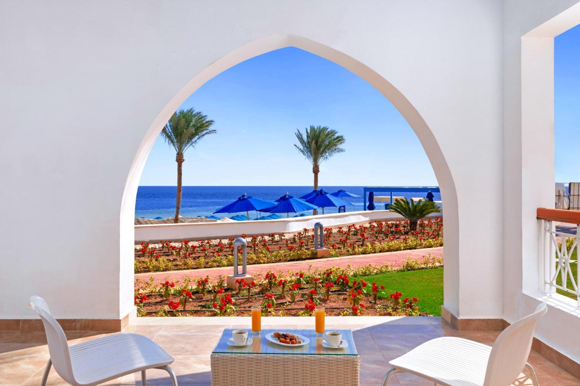 Pickalbatros Palace Sharm - "Aqua Park" Exterior photo The photo depicts a serene view through an architectural archway. In the foreground, there is a small table set with two glasses of orange juice and a plate of snacks. Surrounding the table are two white chairs. Beyond the arch, you can see a calm bl