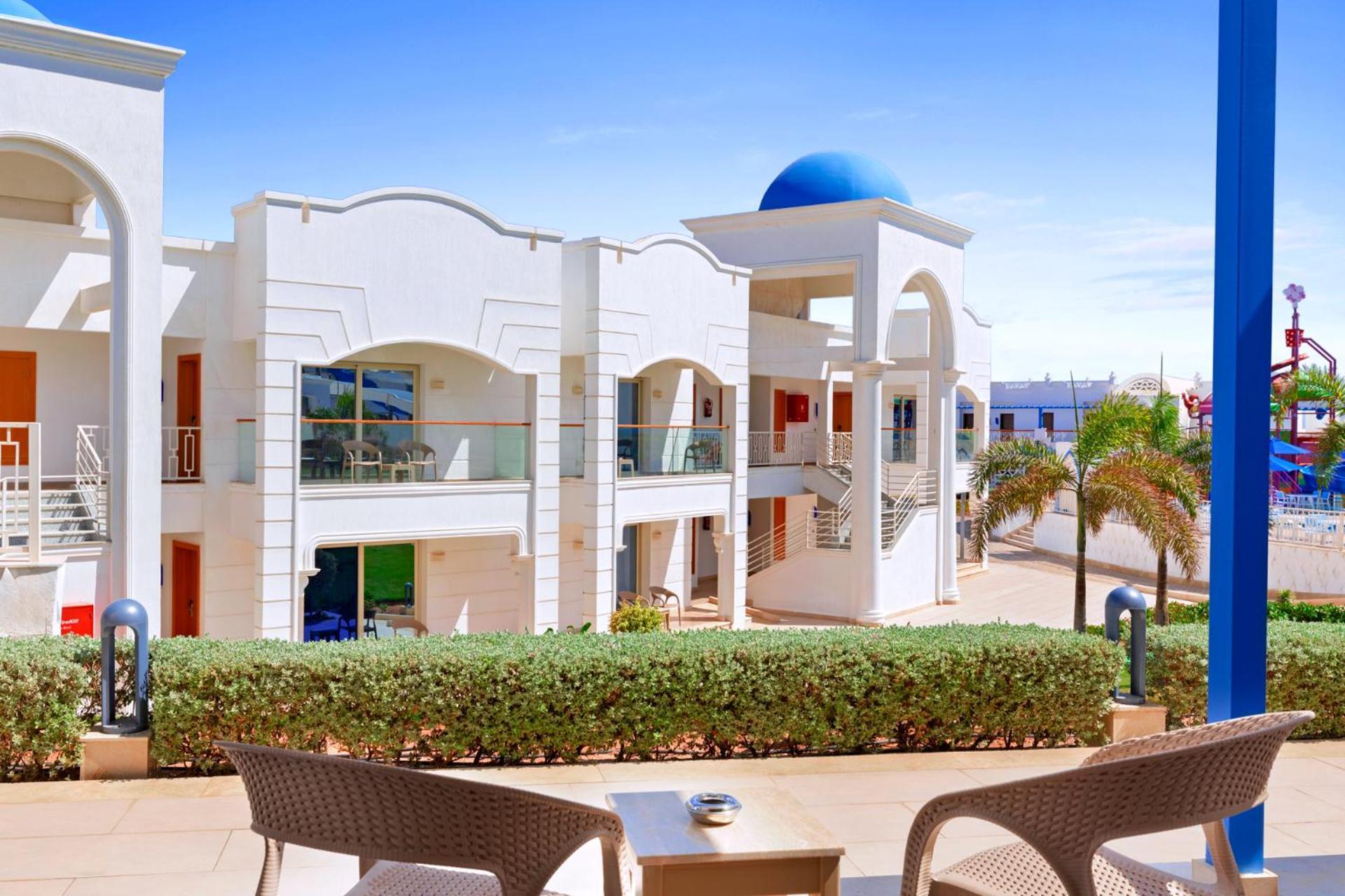 Pickalbatros Palace Sharm - "Aqua Park" Exterior photo The photo shows a modern architectural setting, likely at a resort or hotel. It features white buildings with decorative elements and balconies. The scene is bright with blue skies, and there are palm trees visible, adding a tropical vibe. In the for
