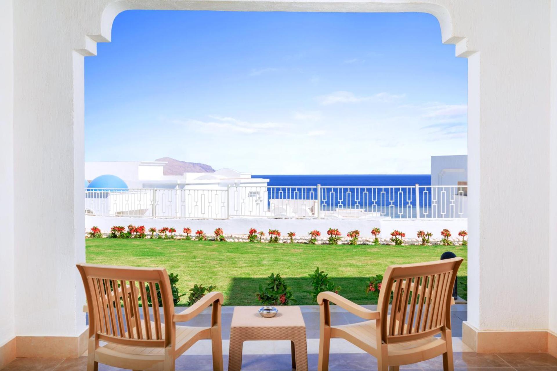 Pickalbatros Palace Sharm - "Aqua Park" Exterior photo The photo shows a scenic view from a terrace or balcony. In the foreground, there are two wooden chairs positioned around a small table. On the table, there is a decorative plant or vase. Beyond the seating area, a well-manicured lawn is visible, ado