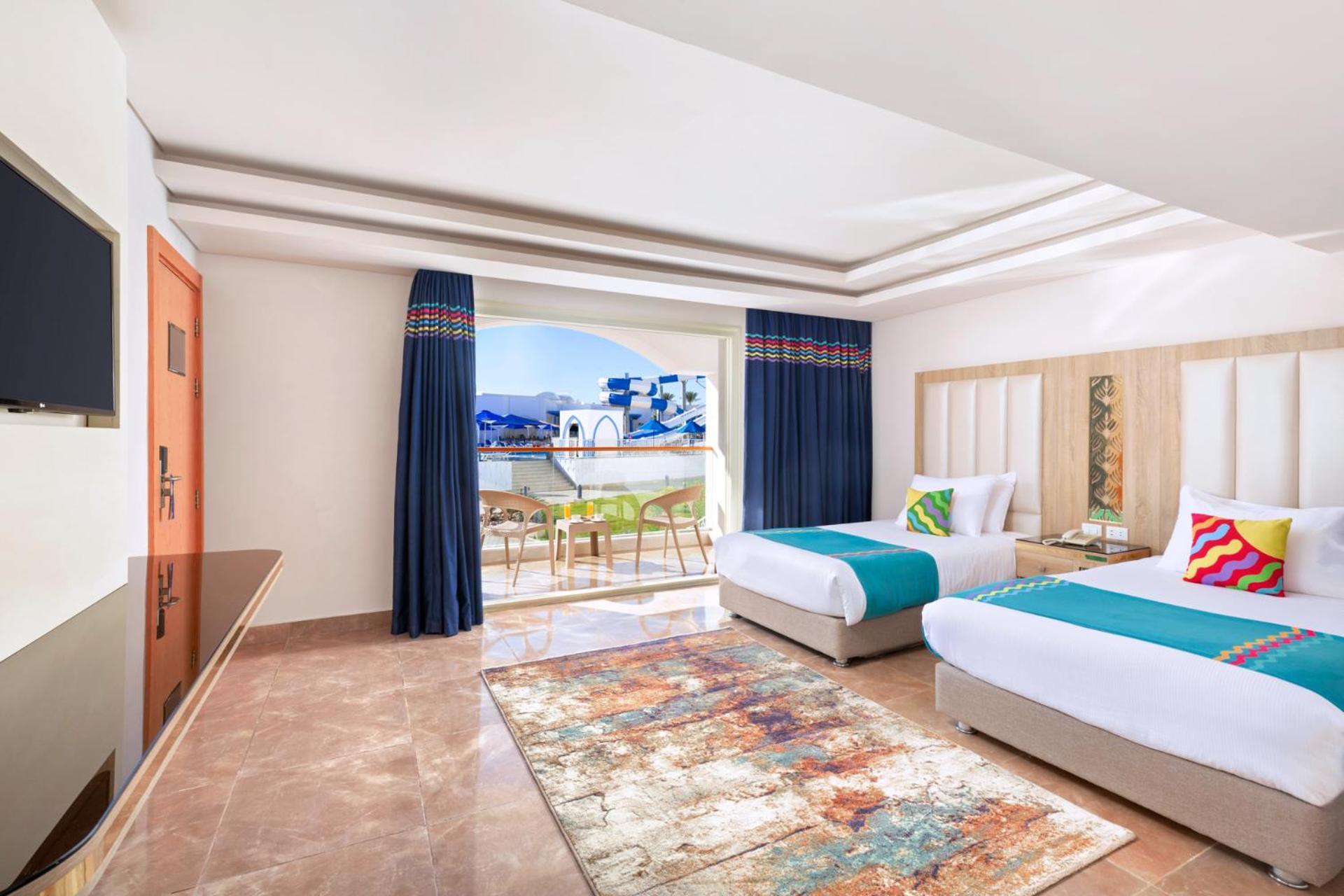 Pickalbatros Palace Sharm - "Aqua Park" Exterior photo The photo shows a well-decorated hotel room featuring two beds, each with colorful accent pillows. The walls are painted in a light color, and there is a large window with blue curtains that offers a view of an outdoor area. A small seating area with