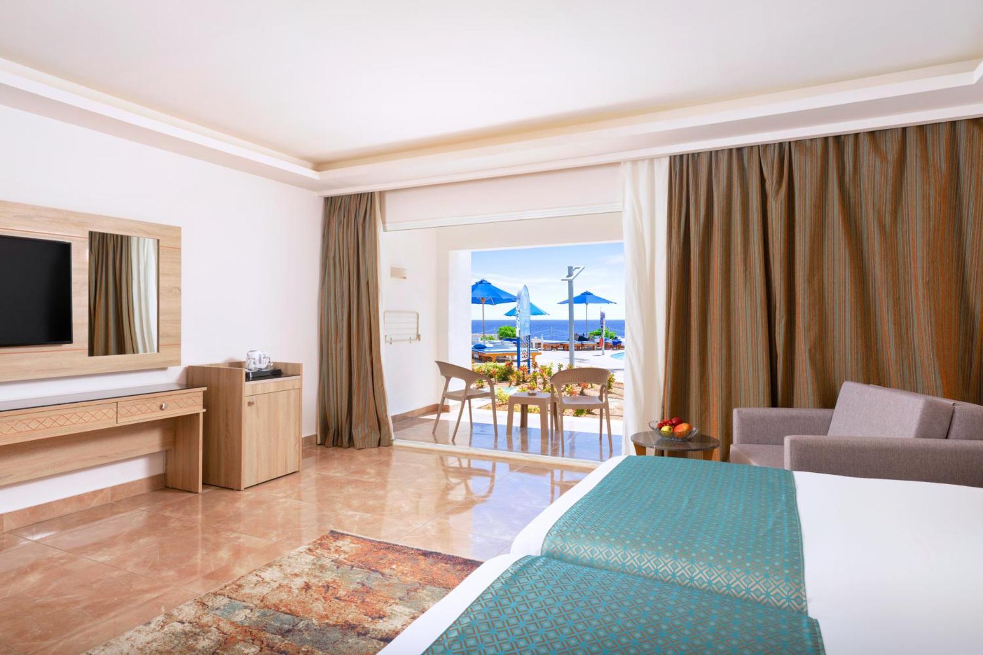 Pickalbatros Palace Sharm - "Aqua Park" Exterior photo The photo shows a spacious hotel room designed with a modern and elegant style. It features two beds with teal-colored bedding, a large mirror, and a television on the wall. There are wooden furniture pieces, including a dresser and a small table. To
