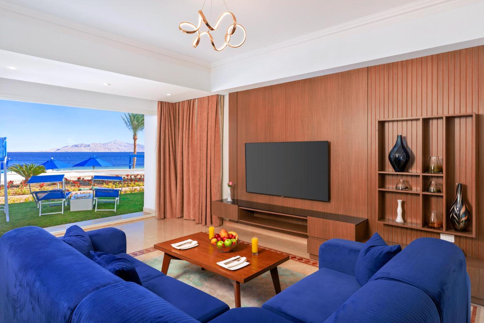 Pickalbatros Palace Sharm - "Aqua Park" Exterior photo The photo shows a modern living room with a stylish design. It features a blue sofa arranged around a wooden coffee table, which has some decorative items, including fruit and candles. A large flat-screen TV is mounted on a wooden wall panel. There a