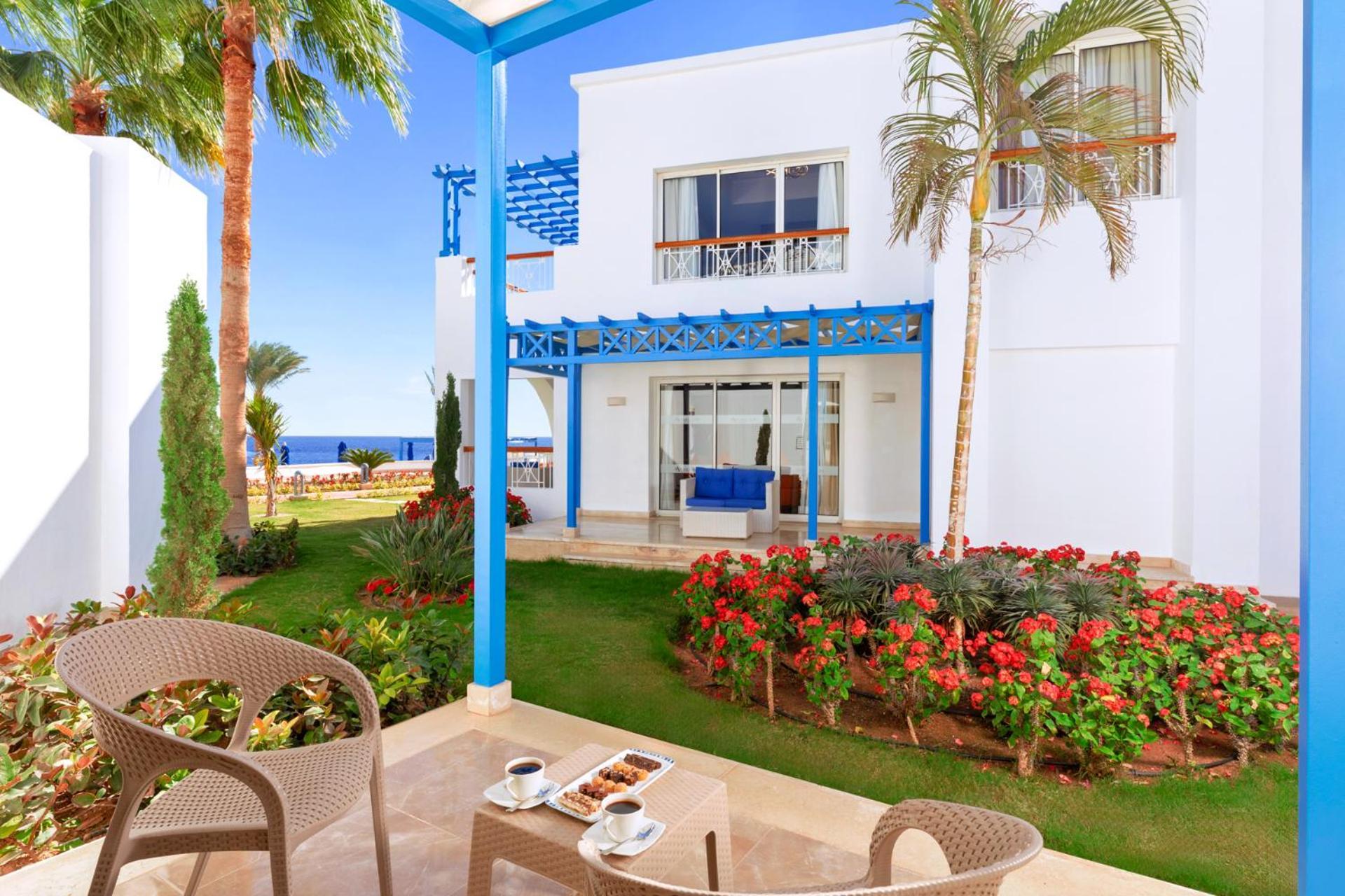 Pickalbatros Palace Sharm - "Aqua Park" Exterior photo The photo shows a picturesque outdoor setting, likely part of a resort or hotel. In the foreground, there are two stylish chairs and a small table with two cups, possibly of coffee or tea, along with a plate of snacks. The background features a well-