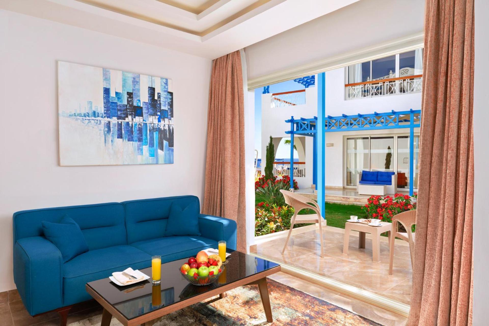 Pickalbatros Palace Sharm - "Aqua Park" Exterior photo The photo shows a bright and modern living space. There is a blue couch facing a glass door that leads to a garden area. In front of the couch, there's a dark coffee table with some fruit and candles on it. A large piece of abstract artwork in blues 