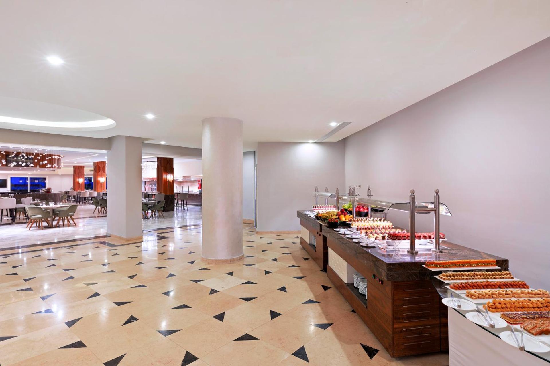 Pickalbatros Palace Sharm - "Aqua Park" Exterior photo The photo shows a modern dining area, likely a buffet or restaurant space. In the foreground, there's a wooden buffet table displaying various food items, including desserts. The floor has a decorative tile pattern, featuring a mix of light and darke