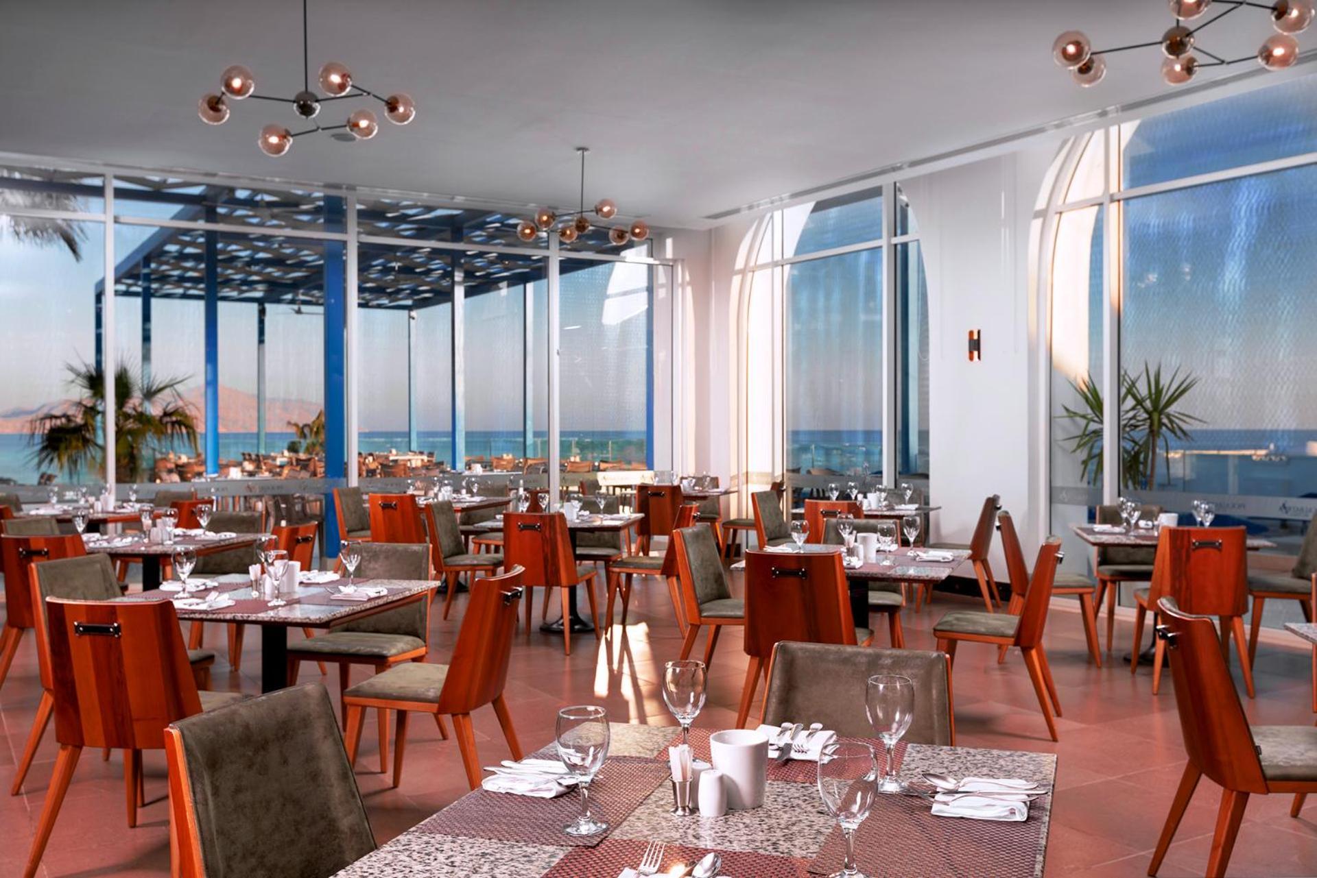 Pickalbatros Palace Sharm - "Aqua Park" Exterior photo The photo shows a modern restaurant interior featuring multiple dining tables arranged neatly across the space. The tables are set with utensils, glasses, and decorations, indicating readiness for guests. Large windows allow plenty of natural light t